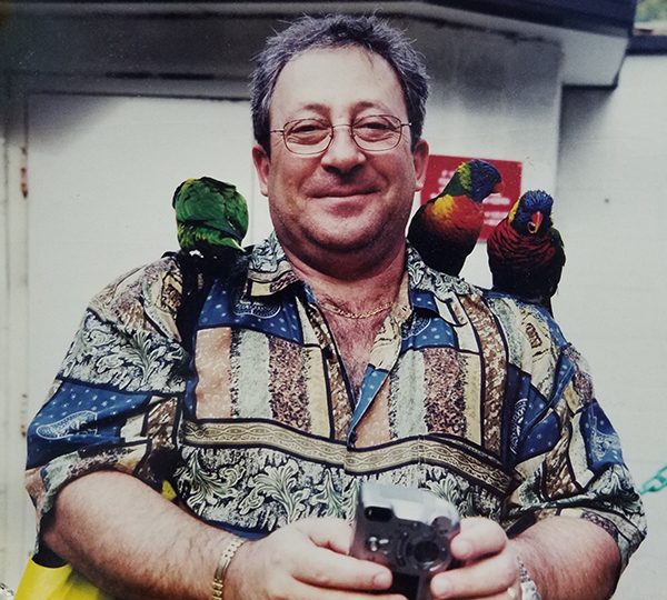 Edward, the founder, smiling in a patterned shirt with three parrots perched on his shoulders, holding a camera. A scanned print.
